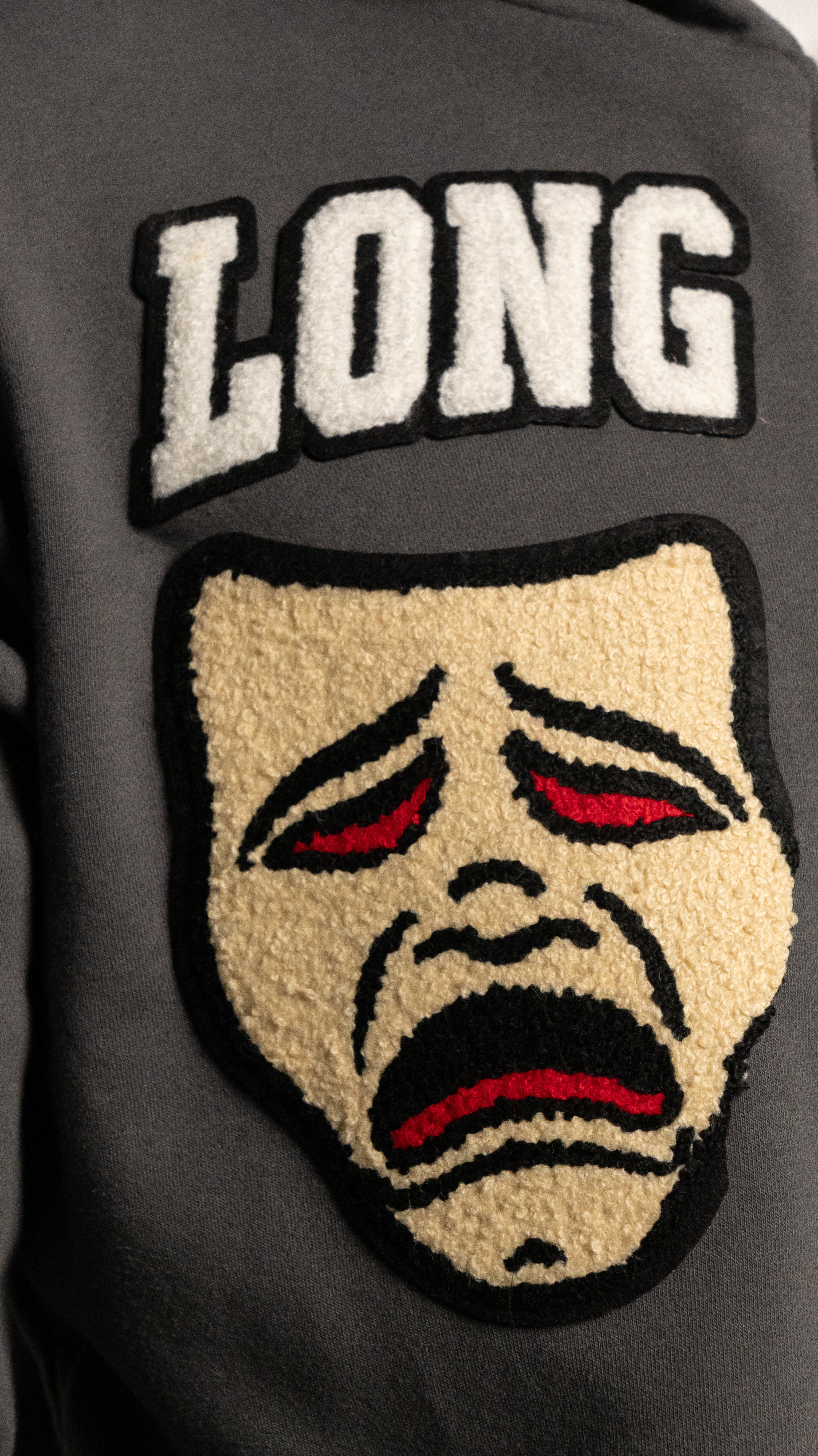 Gray jacket with a patch featuring a face with red eyes and mouth, and the word 'LONG' above it.